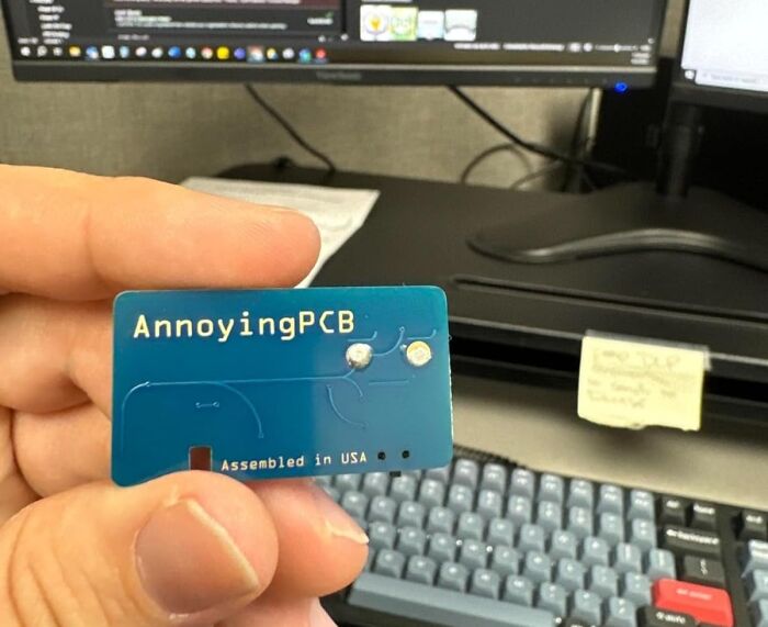 Hand holding an AnnoyingPCB in an office setting for a harmless April Fools prank.