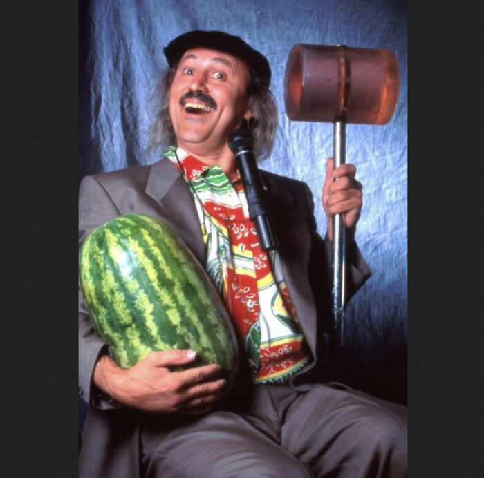 Man with mustache smiling, holding a large watermelon and a mallet, a nostalgic item for creaking joints reminders.