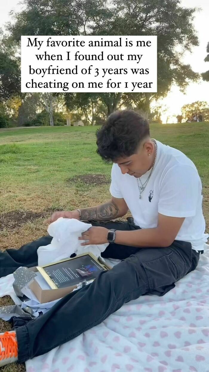 Young man sitting on a blanket outdoors, opening a gift with a relatable meme text about betrayal and cheating.