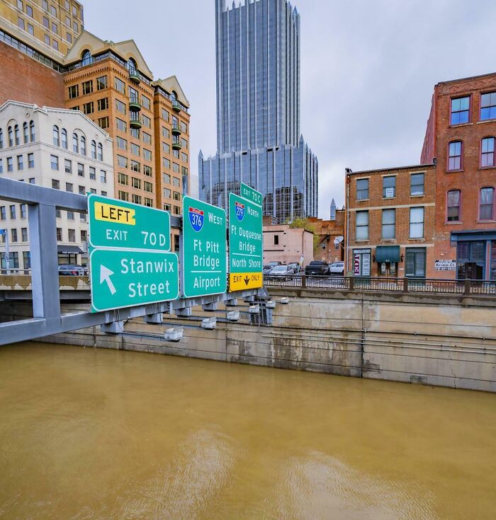 Urban highway signs over a flooded waterway in a cityscape, illustrating wild natural flooding disrupting infrastructure.