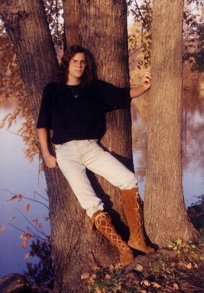 Teenager in cringy childhood photo wearing tall fringe boots and glasses, leaning against a tree near a lake in autumn.