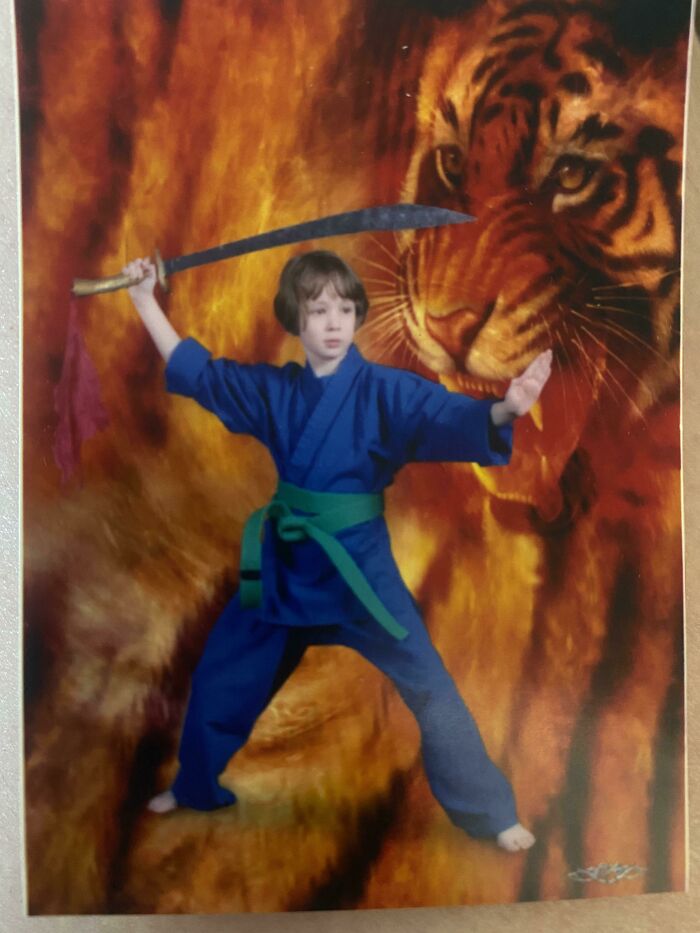Child in blue karate uniform with green belt holding a sword in front of a large tiger backdrop cringy childhood photo.