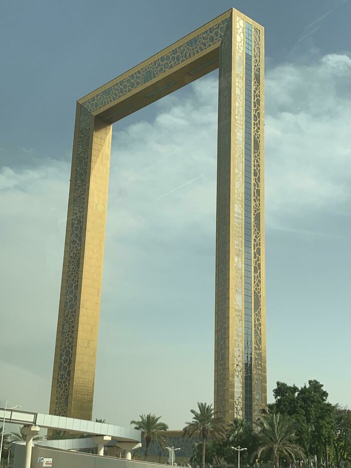Bizarre building shaped like a giant golden frame towering against a partly cloudy sky with palm trees below.