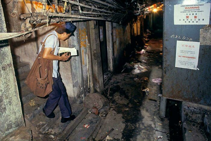 Man in an urban alley holding a book, surrounded by pipes and warning signs, depicting a challenging living environment.