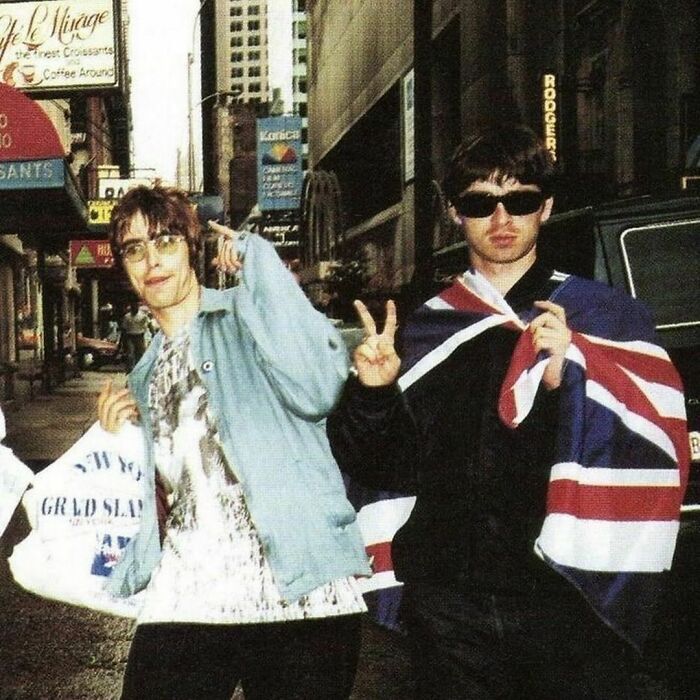 '90s moment with two people in city street, one in a light jacket holding bags, the other draped in a Union Jack.