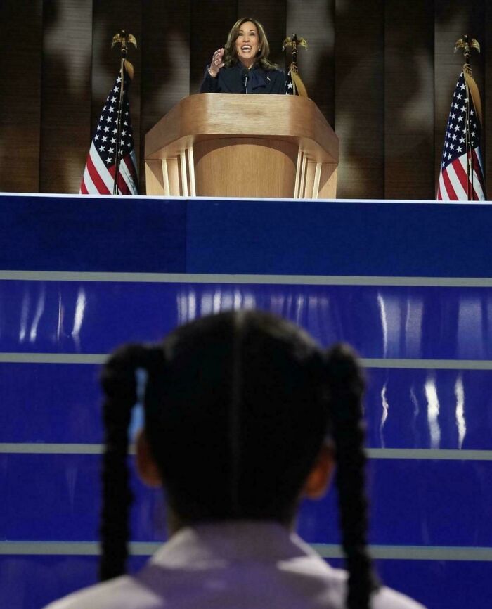 A young girl with pigtails watches a woman speak at a podium, conveying a wholesome moment of support and inspiration.