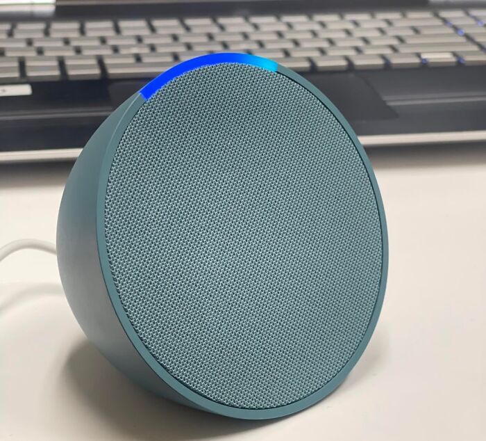 Smart home speaker device with a blue mesh front, positioned near a laptop on a desk.