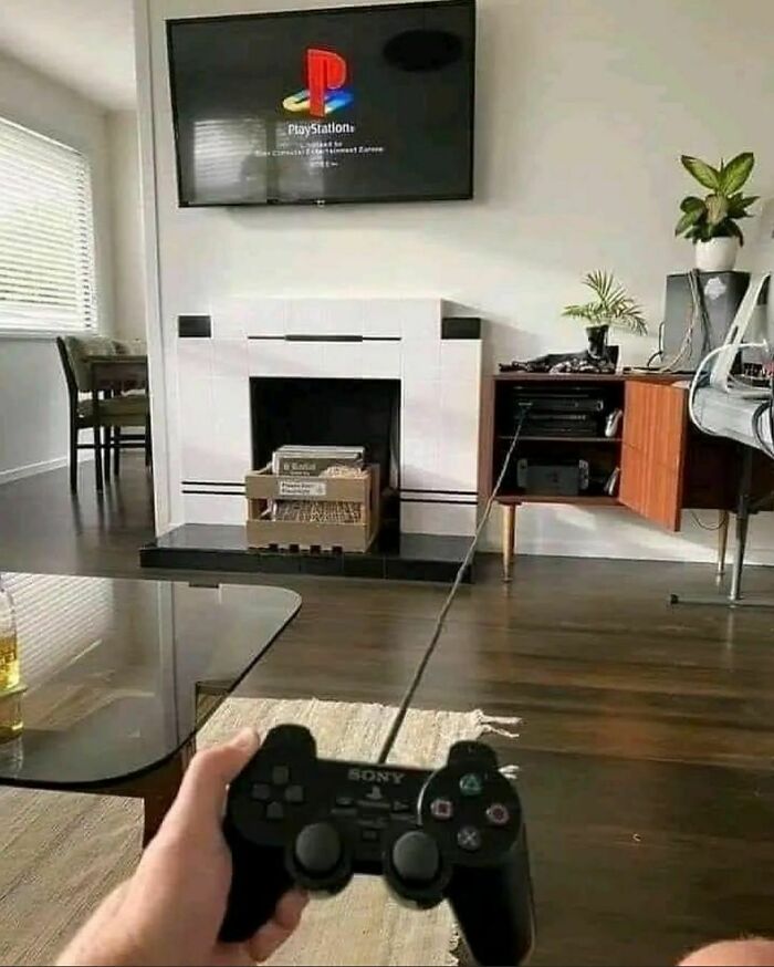 Person holding a Sony PlayStation controller in a living room, TV displaying the PlayStation startup screen.