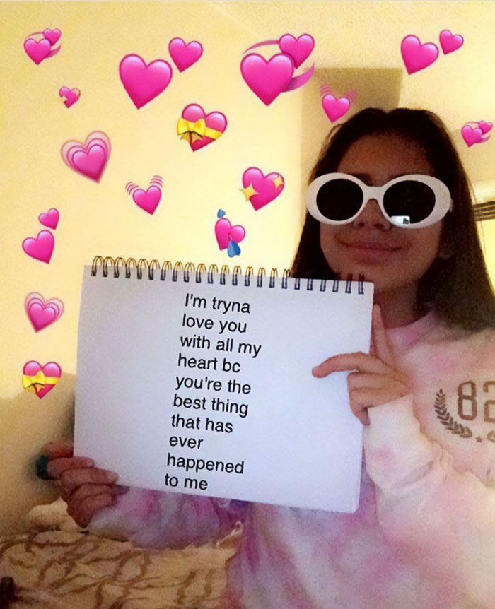  A love meme featuring a girl wearing white sunglasses and holding a notebook. The notebook has a message written on it: "I'm tryna love you with all my heart bc you're the best thing that has ever happened to me." The background is filled with pink heart emojis. The girl is smiling and the setting appears to be indoors.