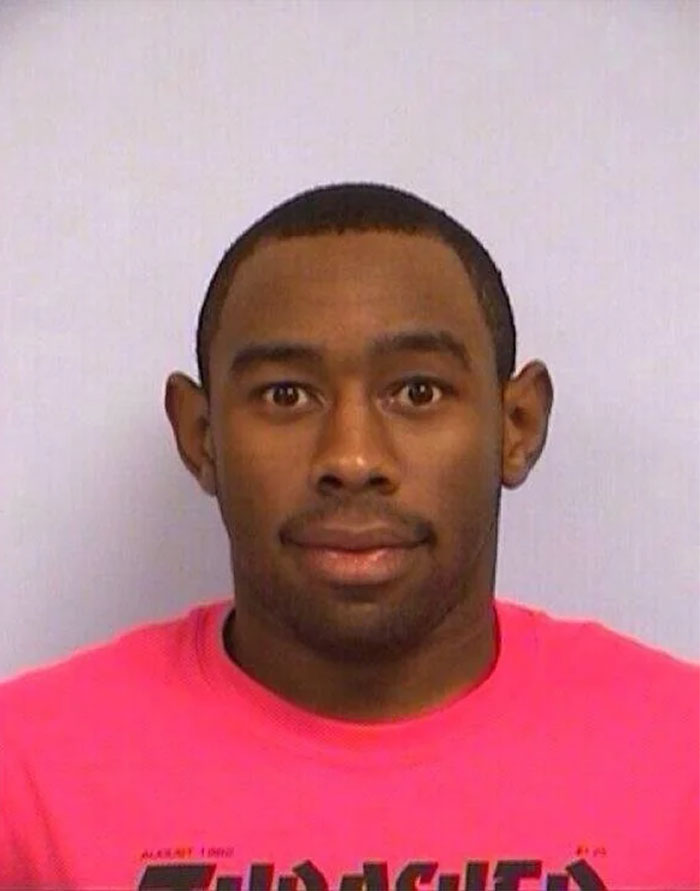 Tyler The Creator mugshot, wearing a pink shirt, with a neutral expression, looking at the camera.