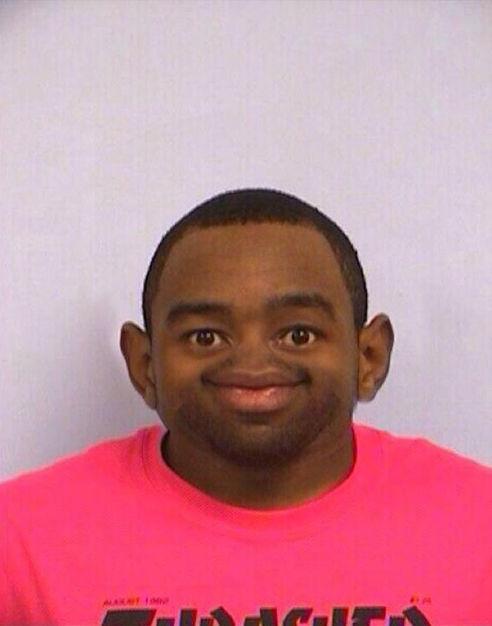 Distorted mugshot of Tyler The Creator, with exaggerated features, wearing a pink shirt and smiling.