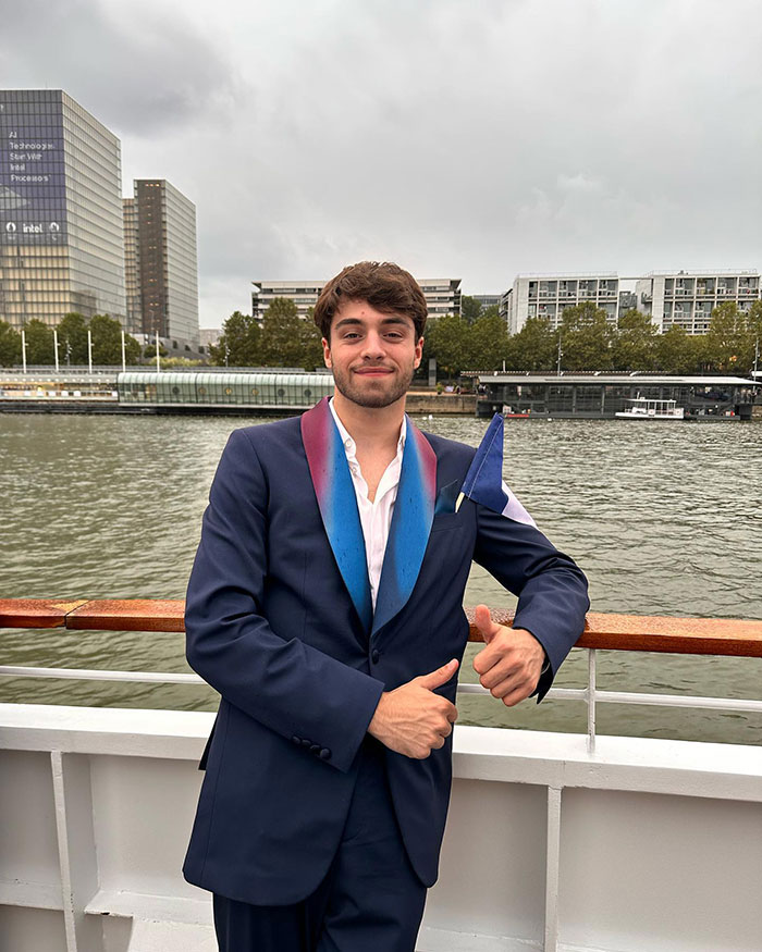 A man in a blue suit giving a thumbs up on a boat, with city buildings in the background. A man in a blue suit giving a thumbs up on a boat, with city buildings in the background.