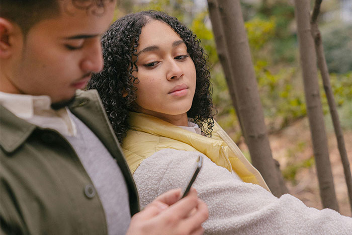 Young woman looking skeptically at man’s phone outdoors, illustrating a scene linked to helicopter parenting concerns.