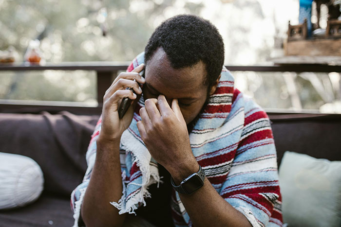 Stressed man covered with a blanket holding phone to his ear, illustrating the pressure of helicopter parenting examples.