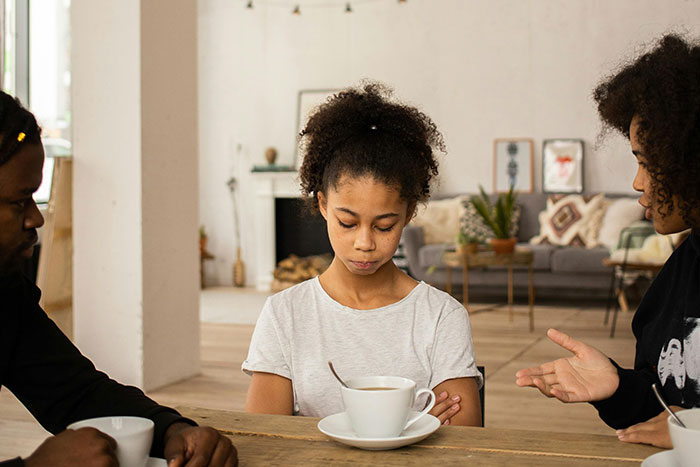 Young girl looking down while two adults around the table show signs of helicopter parenting in a home setting.