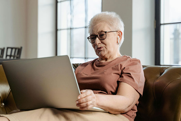 Older woman wearing glasses and earbuds, sitting on a sofa and using a laptop related to helicopter parenting discussion.