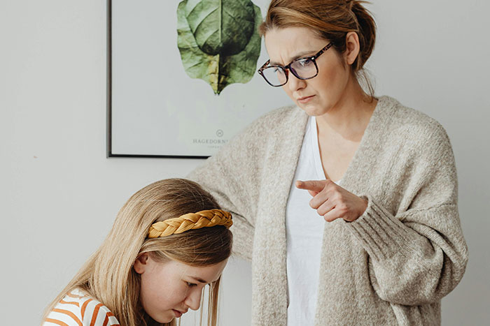 A concerned woman wearing glasses pointing and lecturing a young girl, illustrating helicopter parenting behavior at home.