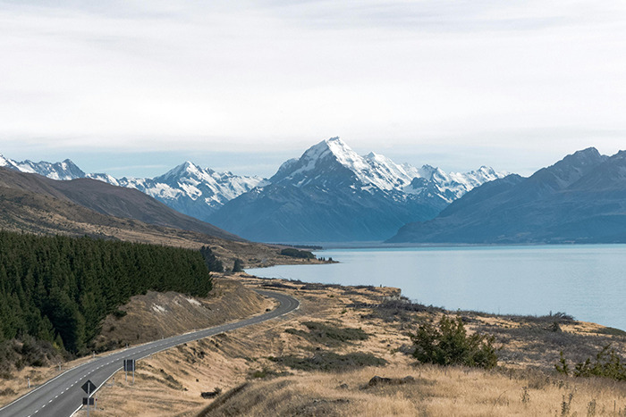 Place visited — winding lakeside road toward snow-capped peaks beside a calm blue lake under a pale overcast sky
