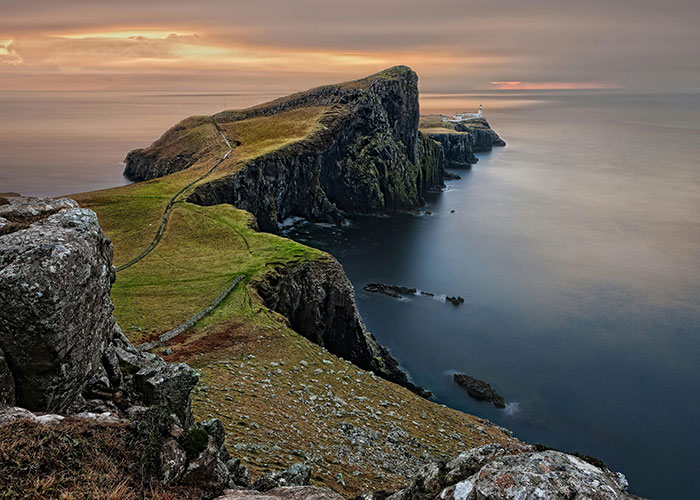 Clifftop coastal place visited with winding grass path leading to a distant lighthouse at sunset over calm sea