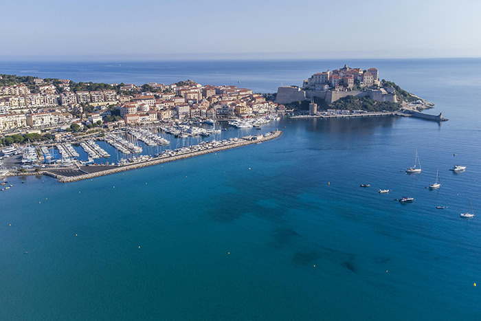 Aerial view of a coastal place visited: marina, sailboats, and a historic fortress on a sunlit Mediterranean shoreline.