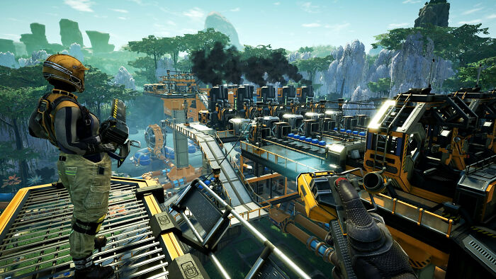 Futuristic game scene with a player overlooking a complex industrial factory in a lush, mountainous environment.