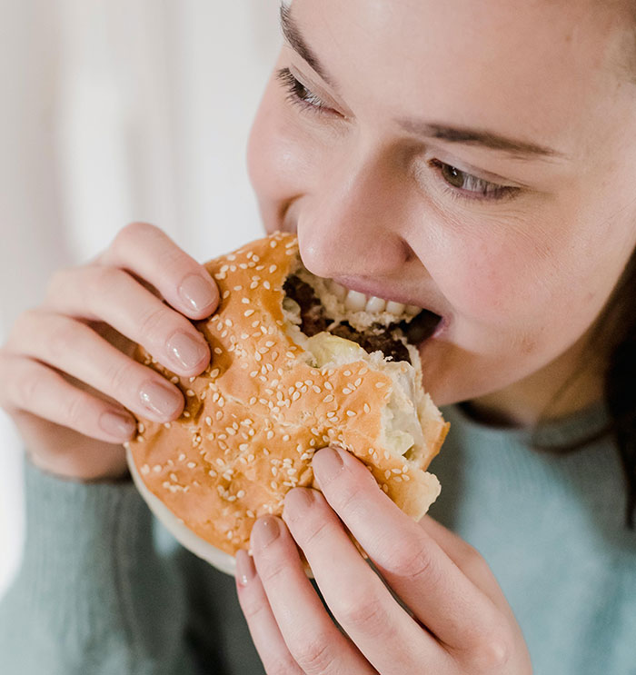 Young woman biting into a sesame burger, illustrating hilarious stereotypes about people’s home countries with humor.