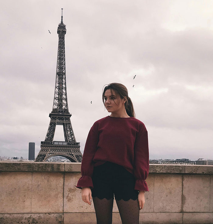 Young woman in a maroon sweater posing near Eiffel Tower, illustrating stereotypes about people’s home countries and cultures.