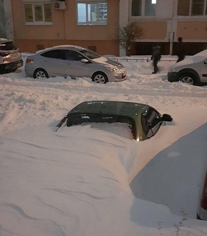 Car almost completely covered in snow in a snowy urban parking area showing nature’s impact on vehicles.