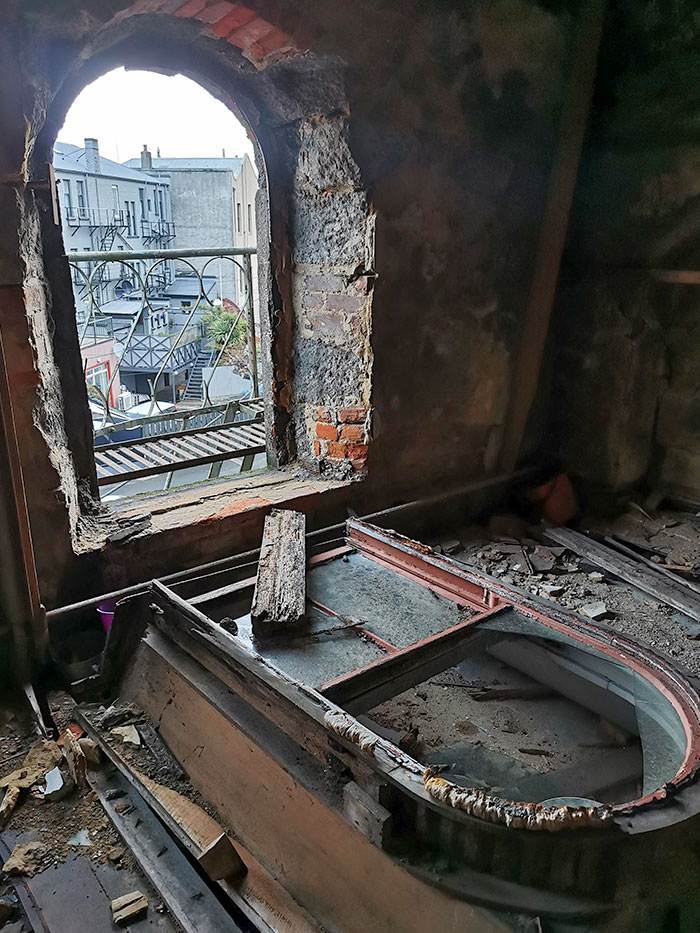 Abandoned room with broken furniture and arched window showing old buildings and nature reclaiming the space.