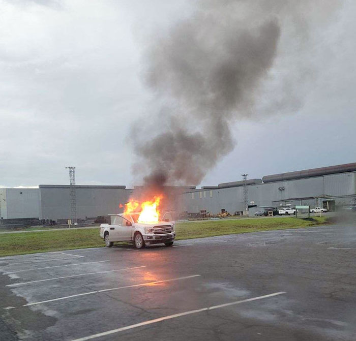 Pickup truck on fire in an empty parking lot with thick smoke rising under cloudy sky showcasing wild natural events.