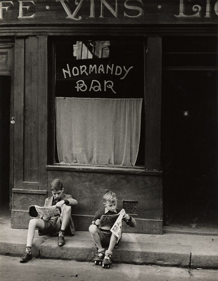 Two boys sitting outside Normandy Bar, reading newspapers in a historical and vintage photo scene.