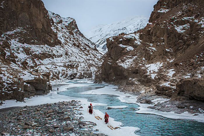 Frozen river flowing through snowy mountains with travelers walking on ice, a top travel photography award-winning image 2024.