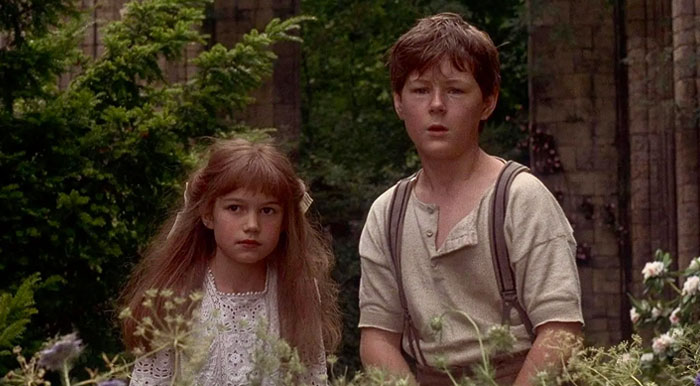 A young boy and girl from “The Secret Garden” movie stand outdoors in a lush garden, looking ahead with curious and slightly concerned expressions. The girl wears a white lace dress, and the boy is dressed in a simple shirt with suspenders. They are surrounded by greenery and flowers, with an old stone structure visible in the background. The scene conveys a sense of mystery and adventure, reminiscent of a period setting. A young boy and girl from “The Secret Garden” movie stand outdoors in a lush garden, looking ahead with curious and slightly concerned expressions. The girl wears a white lace dress, and the boy is dressed in a simple shirt with suspenders. They are surrounded by greenery and flowers, with an old stone structure visible in the background. The scene conveys a sense of mystery and adventure, reminiscent of a period setting.