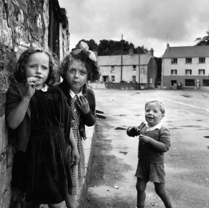 Three children in vintage clothing by a stone wall in a historical black and white photo from a vintage photos collection.