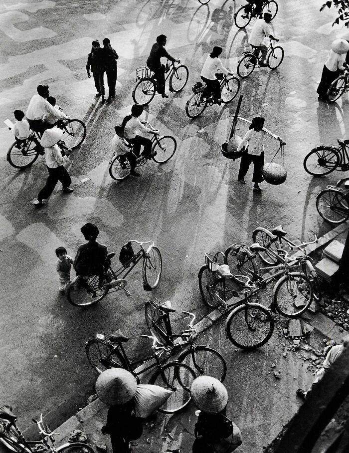 Vintage black and white photo showing people riding bicycles and walking on a busy street in a historical setting.
