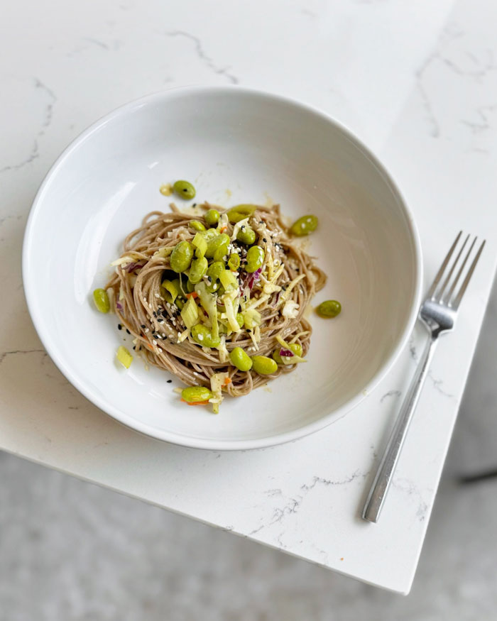 A lunch idea featuring sesame soba noodles topped with edamame, cabbage, and sesame seeds, served in a white bowl on a marble table. A lunch idea featuring sesame soba noodles topped with edamame, cabbage, and sesame seeds, served in a white bowl on a marble table.
