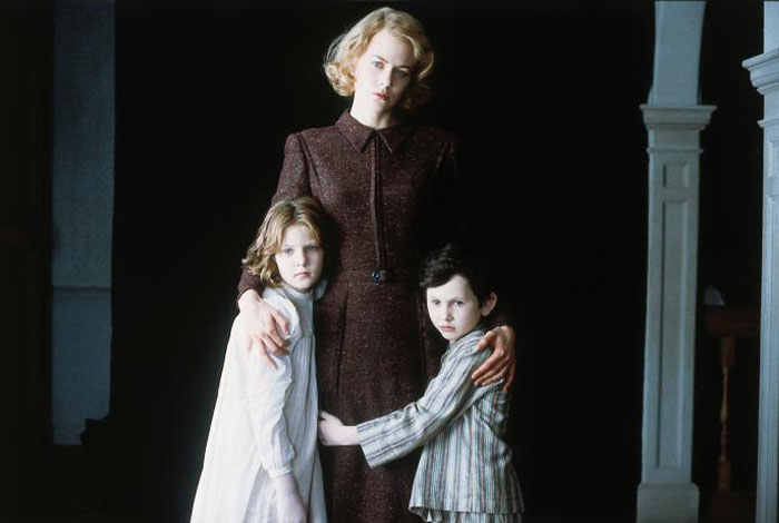 A scene from the movie The Others featuring a woman in a dark, vintage dress standing protectively with her arms around two young children, a girl and a boy. The children cling closely to her, both wearing nightclothes, and have solemn, anxious expressions. The background is dark, creating a haunting and mysterious atmosphere, with the woman looking serious and watchful. A scene from the movie The Others featuring a woman in a dark, vintage dress standing protectively with her arms around two young children, a girl and a boy. The children cling closely to her, both wearing nightclothes, and have solemn, anxious expressions. The background is dark, creating a haunting and mysterious atmosphere, with the woman looking serious and watchful.