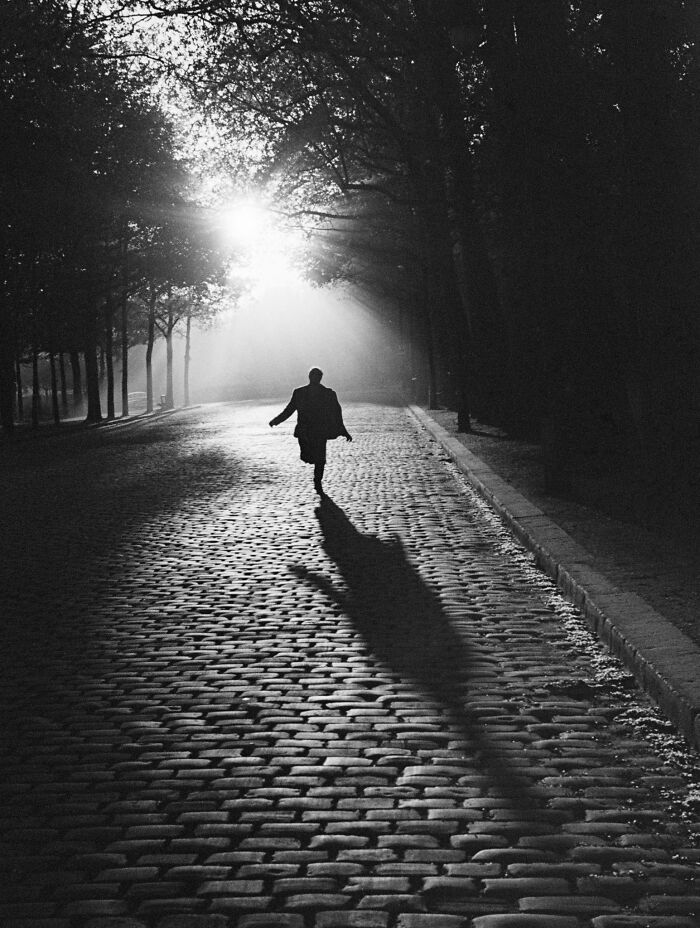 Man walking on a cobblestone street with sunlight filtering through trees in a vintage black and white historical photo.