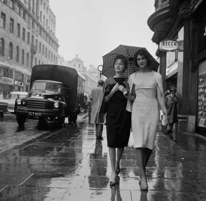 Two women walking on a rainy city street under an umbrella in a vintage black and white historical photo.