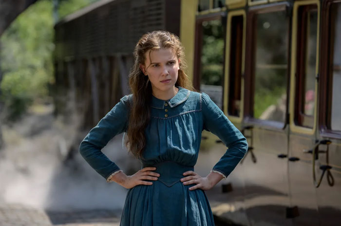 A scene from the movie Enola Holmes featuring a young woman standing with her hands on her hips, wearing a period-style teal dress. She has a determined expression on her face as she stands in front of a steam-emitting train. The setting appears to be an outdoor train station, with greenery in the background, adding to the historical and adventurous tone of the scene. A scene from the movie Enola Holmes featuring a young woman standing with her hands on her hips, wearing a period-style teal dress. She has a determined expression on her face as she stands in front of a steam-emitting train. The setting appears to be an outdoor train station, with greenery in the background, adding to the historical and adventurous tone of the scene.