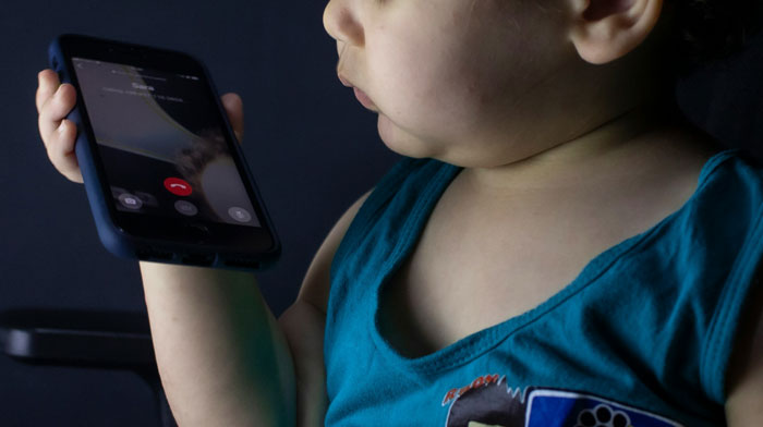 Toddler holding a smartphone close to their mouth, about to listen to or record disturbing voicemails indoors.