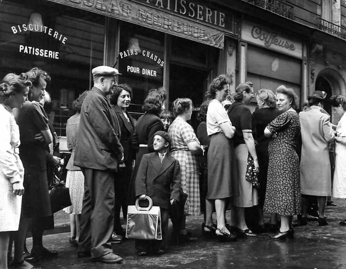 People standing in line outside a bakery, sharing a moment captured in a historical and vintage photo.