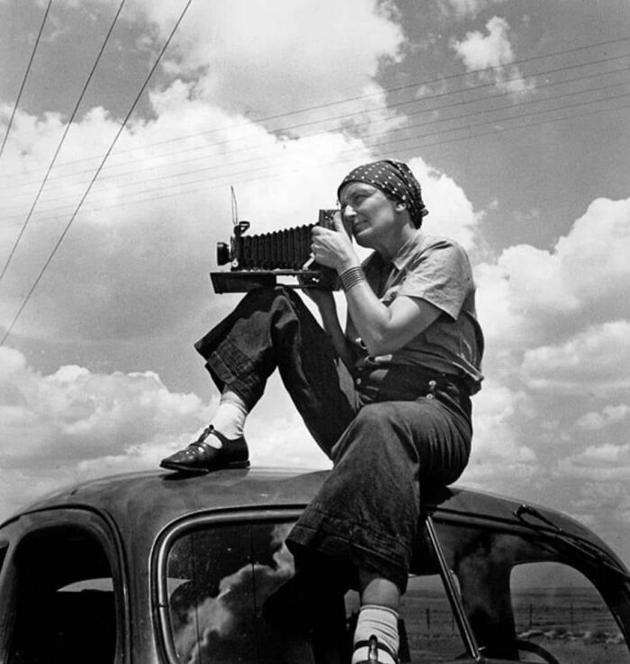 Woman sitting on vintage car roof using old camera outdoors under a cloudy sky, historical and vintage photo moment.