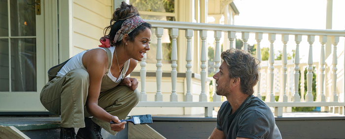 A scene from the movie Falling Inn Love featuring a woman and a man working together on a home renovation. The woman, crouching on the steps of a porch, holds a paintbrush and smiles warmly at the man. She is dressed casually in overalls and a headscarf. The man, sitting nearby, looks up at her with a smile, creating a lighthearted and cheerful atmosphere. The backdrop includes the porch of a cozy house, adding to the scene’s sense of charm and teamwork. A scene from the movie Falling Inn Love featuring a woman and a man working together on a home renovation. The woman, crouching on the steps of a porch, holds a paintbrush and smiles warmly at the man. She is dressed casually in overalls and a headscarf. The man, sitting nearby, looks up at her with a smile, creating a lighthearted and cheerful atmosphere. The backdrop includes the porch of a cozy house, adding to the scene’s sense of charm and teamwork.