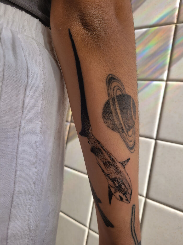 Handpoke tattoo of a shark and planet Saturn on an arm, showcasing intricate black ink design.