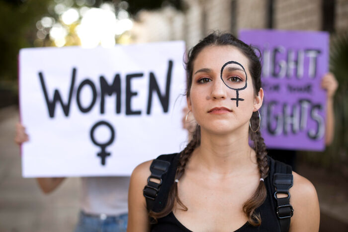 Young woman with feminist symbol on her face, standing confidently in front of signs advocating women's rights.