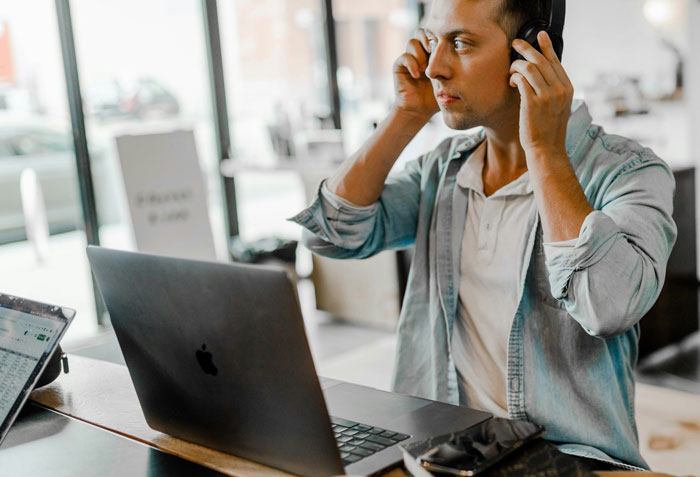 Man putting on headphones while sitting at a table with laptop, reacting to unhinged and disturbing voicemails received.