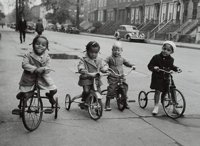 Four children on tricycles on a city street, a vintage historical photo showing childhood moments and old urban life.