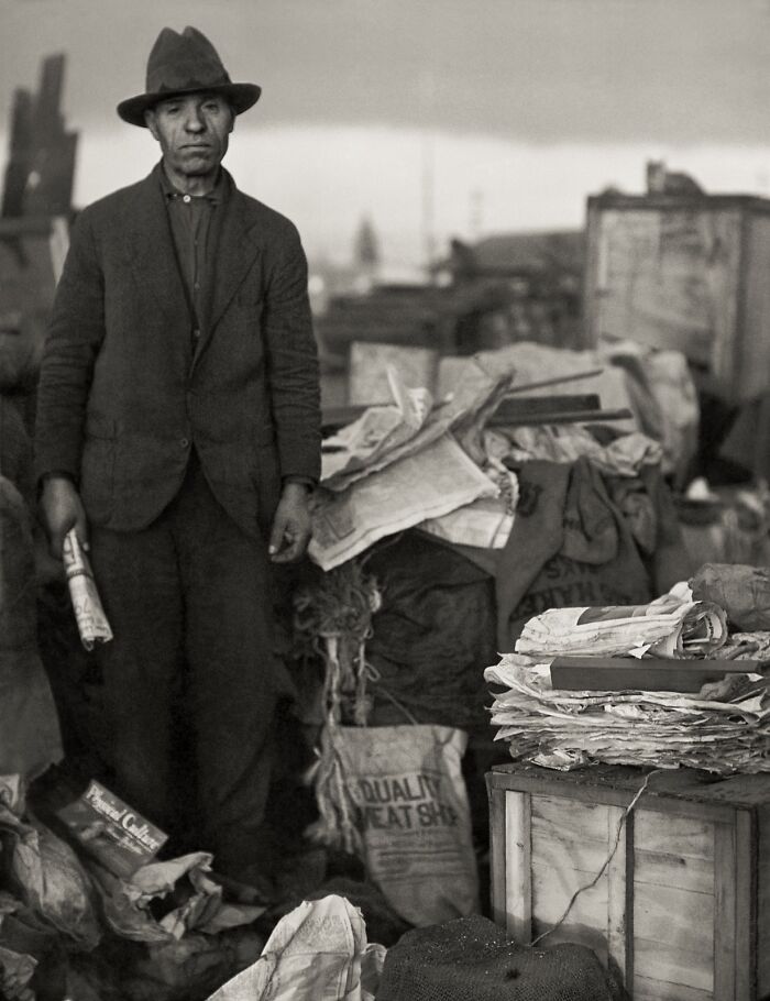 Vintage photo of a man in worn clothes and a hat standing amid piles of paper and sacks in a historical setting.