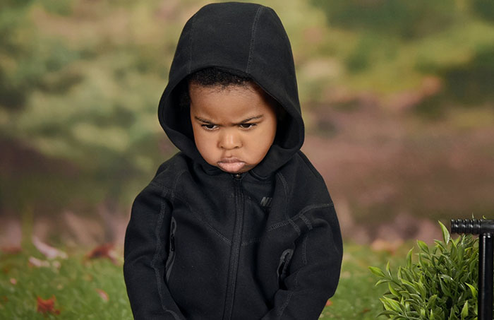 Angry son in first school photo wearing a black hoodie, with a humorous pout, against a blurred outdoor background.